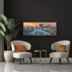 Panoramic mixed-media painting of the Columbus, Ohio skyline at dusk, viewed over the Scioto River with arched bridges and glowing reflections in the water. The artwork features glass and mica sparkle that catches the light, framed in a black floater frame.